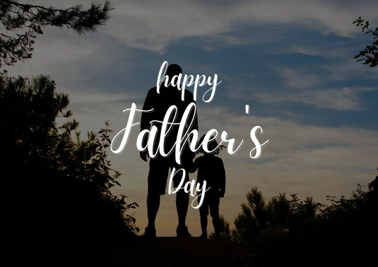 Silhouette of a father and child standing outdoors at sunset, with foliage in the foreground and a blue sky with clouds in the background. In the center, the text "happy Father's Day" appears in elegant white script.