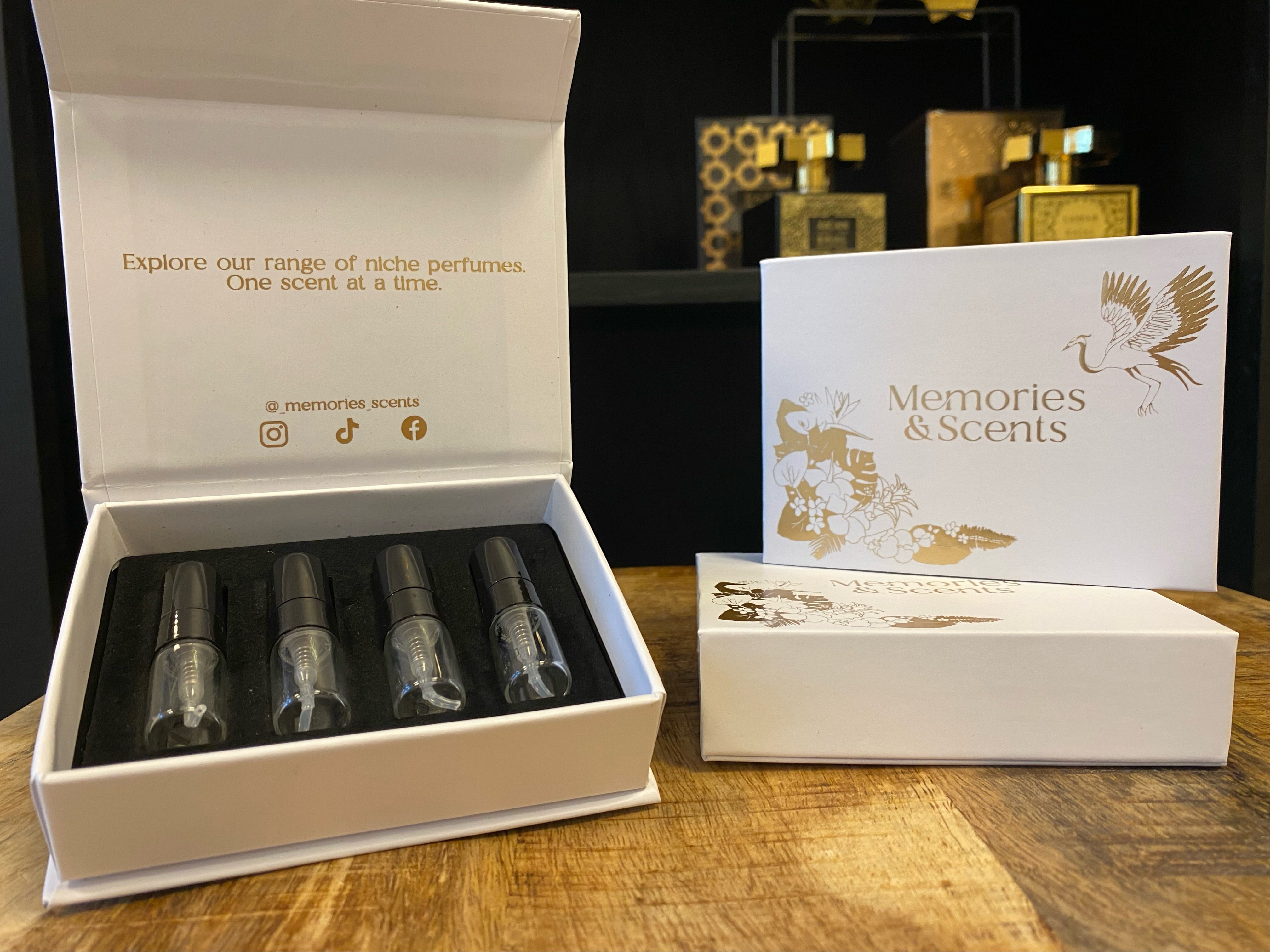 An open 'Memories & Scents Review Box' showcasing four perfume samples in elegant glass vials, set against a stylish background. The box features a gold and white design with the brand name prominently displayed on the exterior.